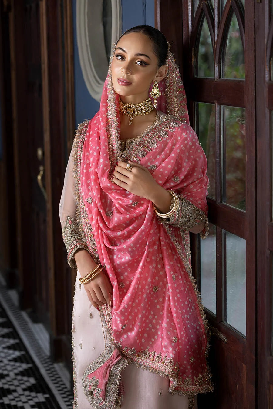 A woman wearing traditional Indian attire, featuring a pink embroidered dupatta and a light beige kurta, standing in front of a dark wooden door.
