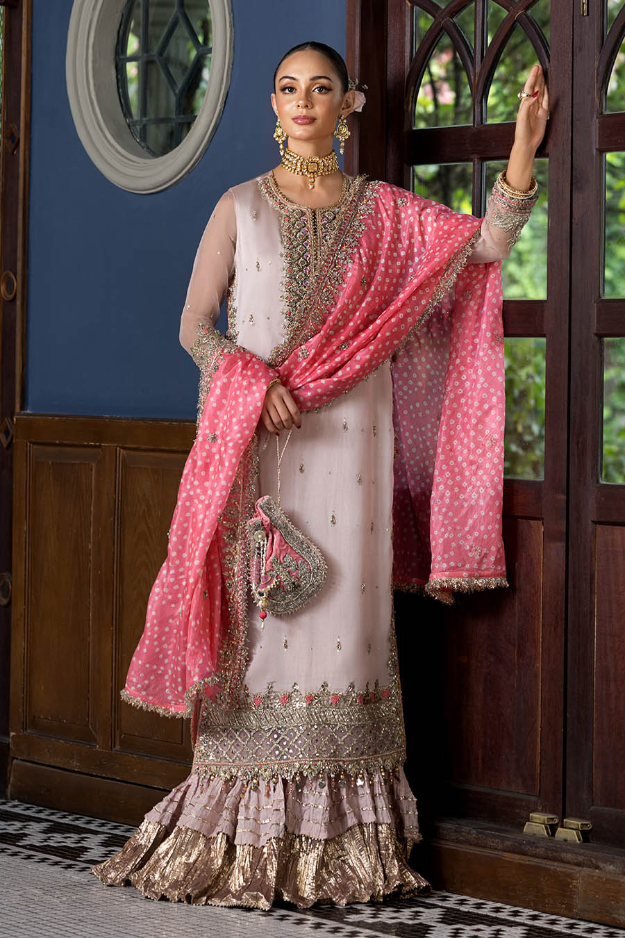 A woman models an elegant traditional South Asian outfit featuring a light pink kurta with intricate gold embroidery, a matching pink dupatta with white polka dots, and a ruffled metallic gold skirt, posed in front of a dark wooden door.