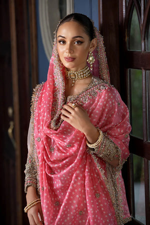 A woman wearing a pink traditional outfit with intricate gold embroidery, a matching embroidered dupatta, and gold jewelry, standing indoors near a dark wooden door.