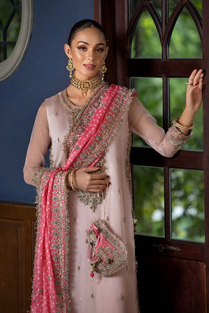 A woman models a traditional Indian outfit consisting of a light pink kurta with heavy silver embroidery, a matching pink dupatta with silver embellishments, and gold jewelry, standing in front of a dark wooden window.