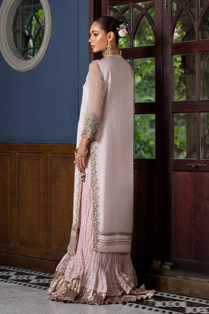 A woman models a traditional three-piece salwar kameez suit in a dusty pink color with gold embroidery, standing in front of a dark wooden door and blue wall.
